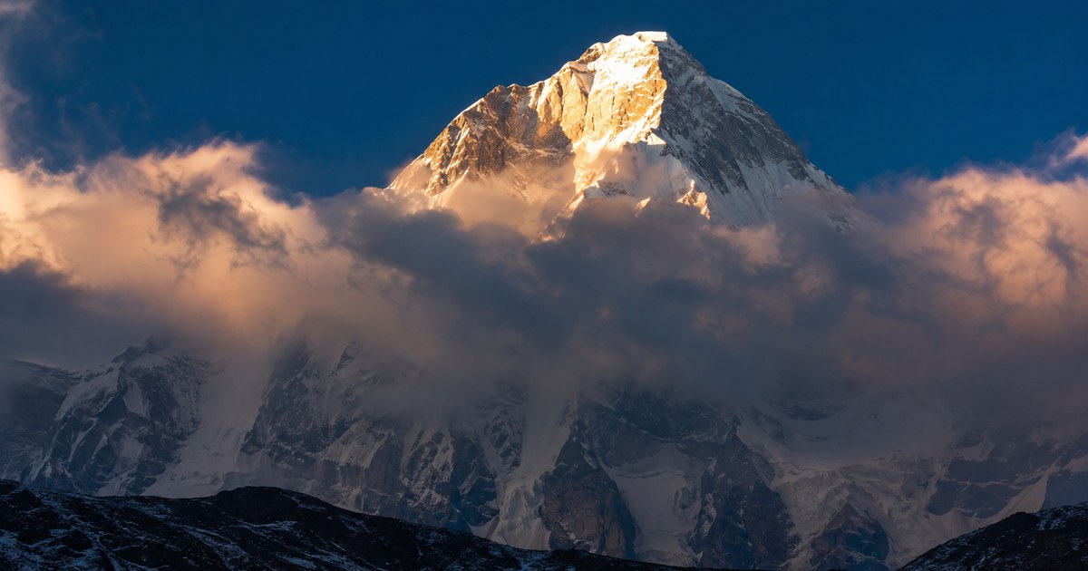 A massive snow-covered mountain rising alone above low tundra and a broken layer of cloud