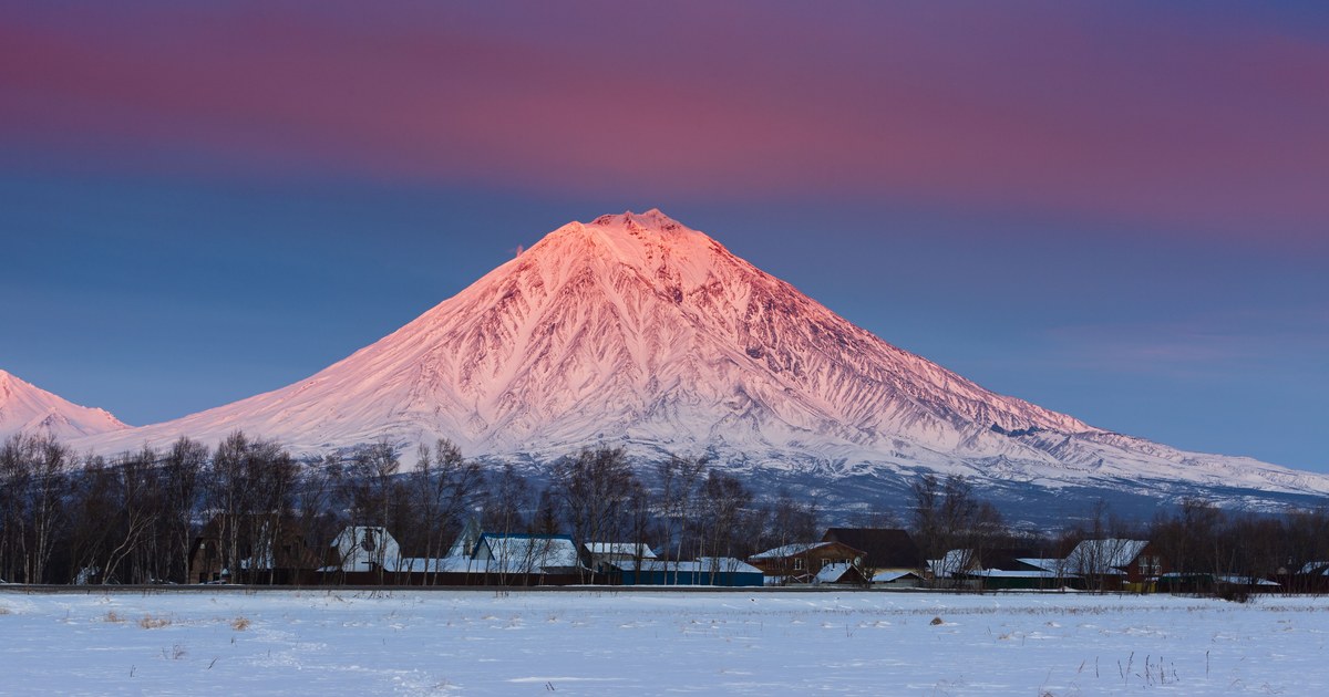Snow-covered volcanic cone catching pink alpenglow above a flat snowy plain