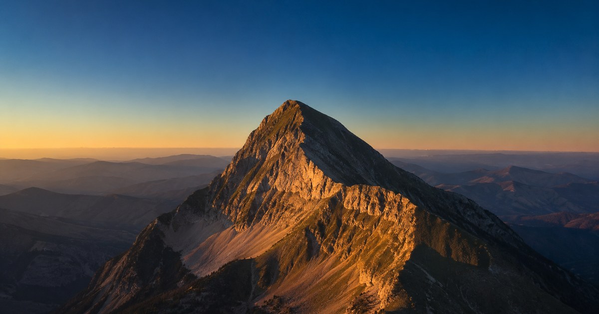 A single snow-capped summit standing far apart from a distant range across a wide, empty plain at dusk